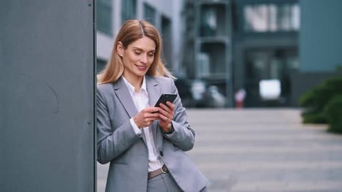 Holding smartphone. Woman in formal clothes is outdoors near the business building