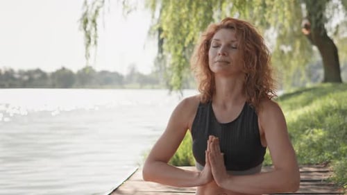 Rothaarige Frau praktiziert Yoga-Meditation im Freien an einem ruhigen Flussufer an einem sonnigen und friedlichen Tag