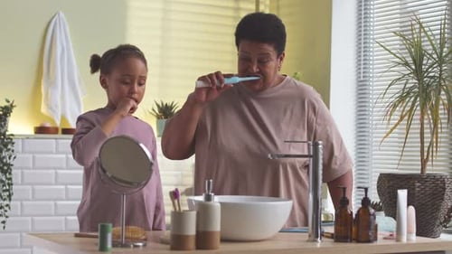 Girl and Adult Brushing Teeth in Bright Bathroom