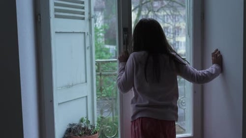 Back of pensive child standing by window staring at view from second floor apartment in moody