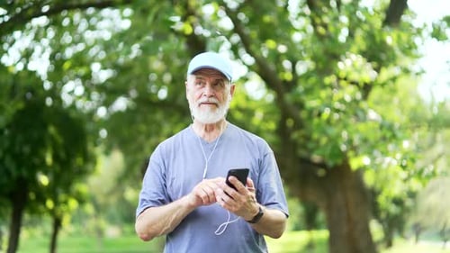 Senior active gray haired bearded man in headphones uses a smartphone while walking in urban city