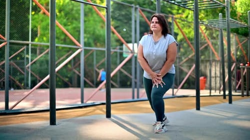 Woman with overweight in gray t-shirt and leggings exercising on a sports field in a park to lose we