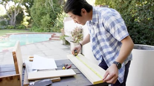 Measuring wooden plank, asian man working on DIY project in backyard workshop