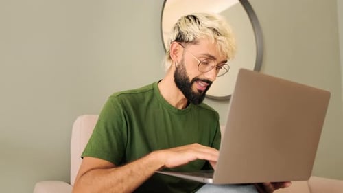 Young blond man with a beard works on a laptop while sitting in a cozy living room