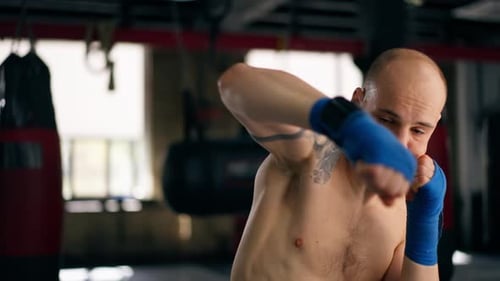 Closeup in a Boxing Fight Club Bald Young Boxer with Tattoos in Blue Bandages Trains Punches Shadow