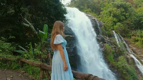 Woman in Blue Dress Admiring Majestic Waterfall in Lush Green Forest