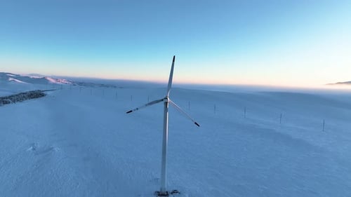 Slow motion of a single wind turbine stands in a vast, snow-covered field under a clear sky at dawn