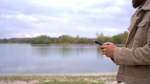 Bearded Man Uses Phone by Lake on Cloudy Day