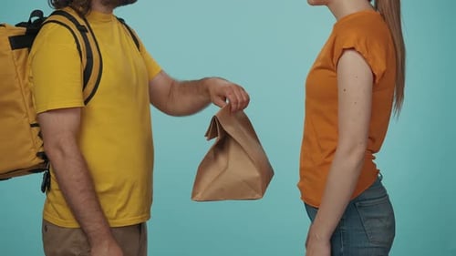 Delivery Man in Yellow Uniform with Portable Backpack Gives a Girl Customer Paper Bag with Food