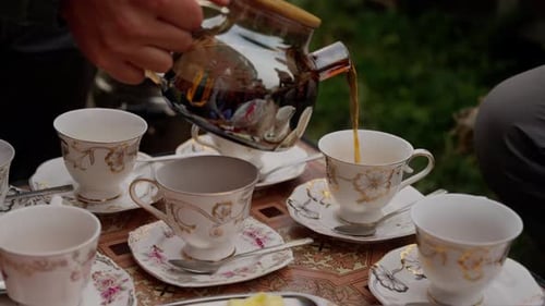 Man in Dark Clothing Pours Tea From a Silver Teapot Into Elegant White and Gold Cups Arranged on a