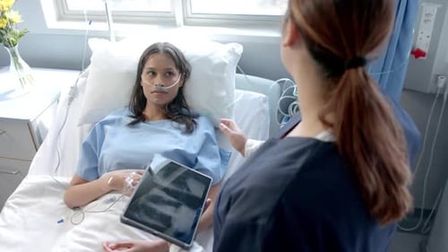 Diverse female patient in hospital bed and female doctor using tablet talking, slow motion