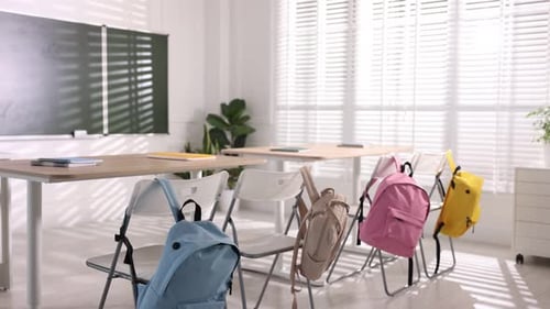 Green chalkboard, desks and chairs with backpacks in classroom. Camera moving left