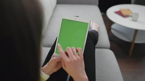 Young Woman Scrolling Modern Digital Tablet Device with Green Screen Monitor Sitting on the Sofa at
