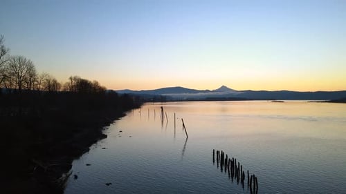 Drone flying low above a large lake during sunrise, trees and mountains on the coast and shore.
