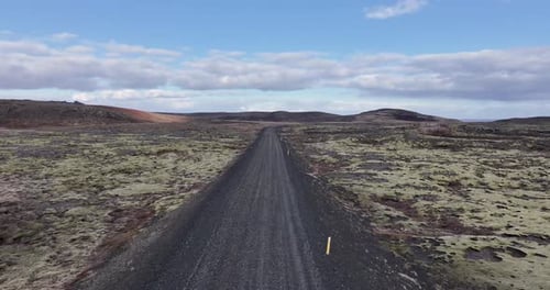 A drone captures a desert road in Iceland under a clear blue sky with fluffy clouds.
