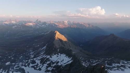 Dramatic drone aerial of snow-flecked alpine ridge at sunset, descending into a deep mountain valley
