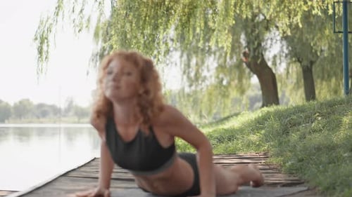 Young Woman Practicing Yoga Outdoors on a Wooden Deck Near a Calm Lake Stretching in the Morning