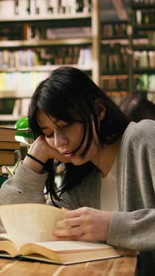 Vertical Video Tired Woman Student Leans on Her Hand While Reading Open Book Under Green Lamp
