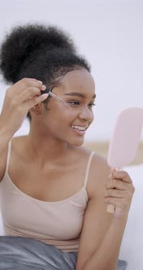 Une adolescente afro-américaine sourit vivement en appliquant du sérum près des yeux à l'aide d'un compte-gouttes pendant que...
