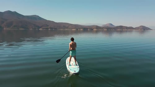 Aerial drone flying over a lake, man doing stand up paddle, with mountains at sunrise. SUP.