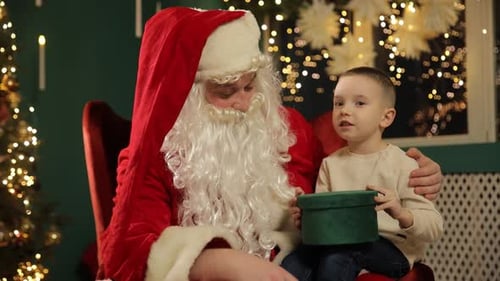 Santa Claus Sits with a Boy Who Holds a Bright Green Christmas Gift Box During a Warm