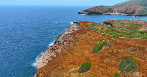 Orange bare rocks with some spots of greenery. Blue waterscape surrounds the rocky shore.