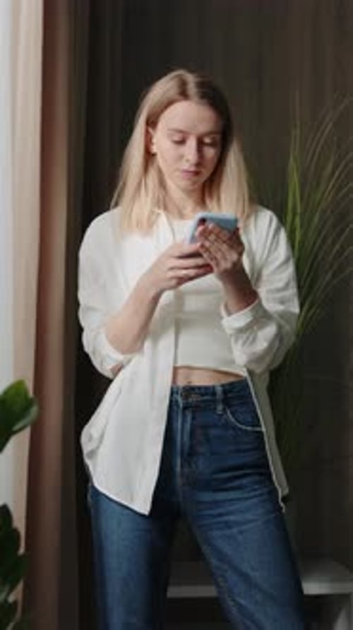 Footage of Thoughtful Young Woman Dressed in Casual Wear Looking Out of Window Holding Smartphone in