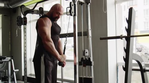 A Young Man Bodybuilder in the Gym Trains His Arms Muscles on a Exercise Machine and Demonstrates