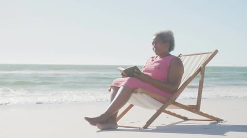 Happy senior african american woman sitting on deck chair and reading book at beach, in slow motion