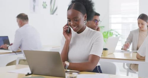 Businesswoman talking on a call on a phone and using laptop in an office with colleagues