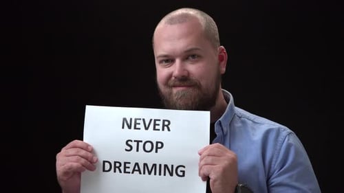 Bald Bearded Man in Blue Shirt on Black Background Shows White Sheet of Paper with Inscription Never