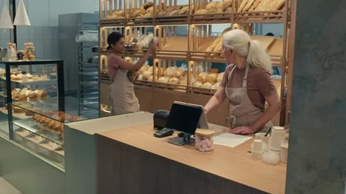 Woman Putting Bread on Shelf and Helping Colleague in Bakery