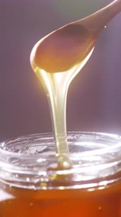 A female hand holds a wooden spoon as golden honey drips gracefully into a glass jar below. Warm lig