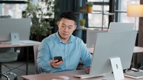Junger Mann arbeitet am Schreibtisch mit Computer und Smartphone in hellen Büroräumen