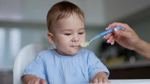 Adorable baby boy eating porridge from spoon. Caring mother giving food to her son. Side view.