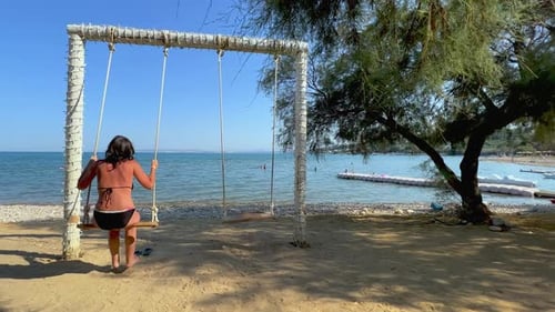Back view of adult woman on summer holiday enjoy swinging on rope swing looking at calm sea