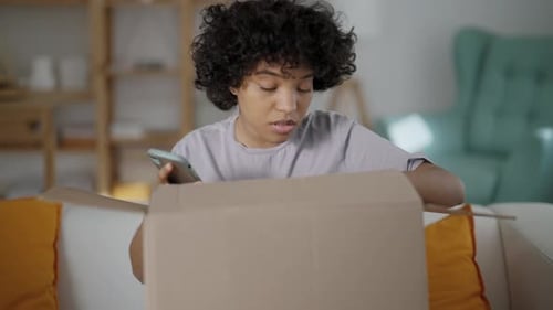 Woman with Phone Opens Cardboard Box at Home