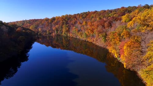 Eine Luftaufnahme eines reflektierenden Sees an einem sonnigen Tag mit bunten Herbstbäumen auf beiden Seiten. Der Tropfen