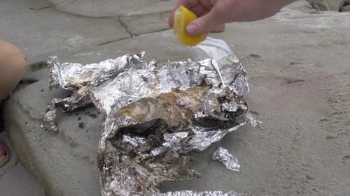 Close up shot of a man pouring lemon over a cooked fish in foil on the beach.