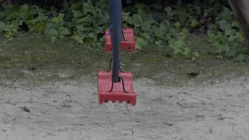 Red Swings With Black Chains Standing Still On Sand Park With Some Plants