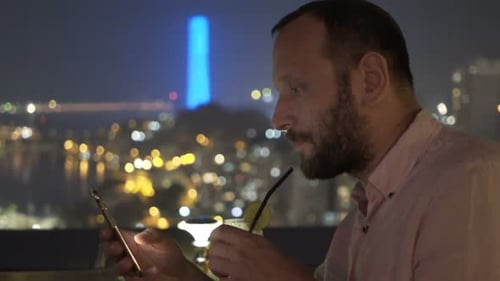 Young Man with Smartphone Drinking Cocktail Sitting on Terrace in Bar At