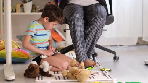 Little Boy Plays Under His Mother's Desk While She Talks Over a Video Conference Working Office at