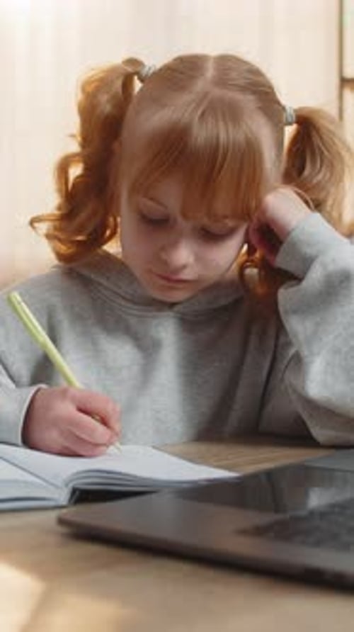 Tired Young Girl Doing Homework at Table with Laptop Writing in Notebook and Showing Lazy Expression