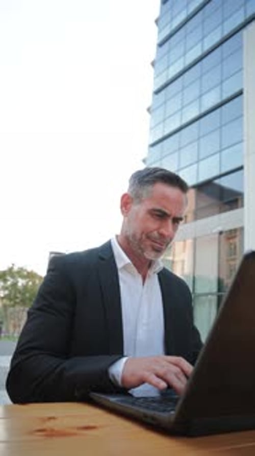 Vertical Focused Mature Businessman Working on Laptop at Outdoor Cafe Engaged in Professional Tasks