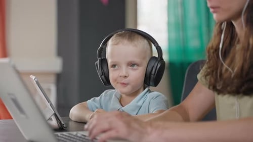 Kind mit kabellosen Kopfhörern benutzt Tablet, wenn seine Mutter zu Hause arbeitet