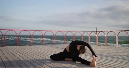 A Calm Brunette Girl in a Black Sports Uniform Does Yoga and Stretching on the Embankment Near the