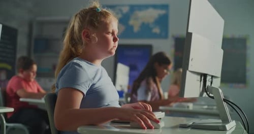 Blonde Girl in Blue Shirt Works on Desktop Computer While Classmates Sit at Desks