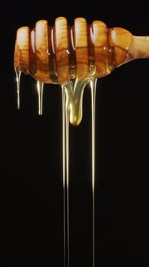 Drops of honey flow from a wooden honey dipper and slowly drip down, black background.