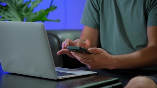 Man Sitting On Couch In Living Room Holding Phone And Using Laptop Computer Shopping Online