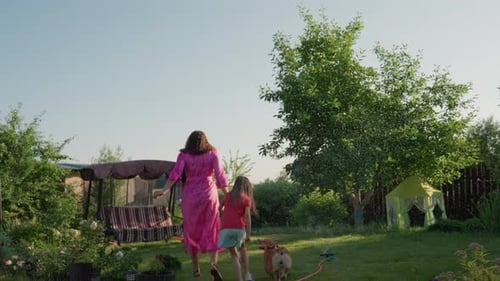 White Mother Walking With Daughter Toward Garden Swing Pink Robe Small Dog At Heel Backyard Hose
